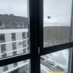 Панорамные окна вдоль лоджии, тёмные рамы; на потолке вентиляционный короб, черновая отделка.